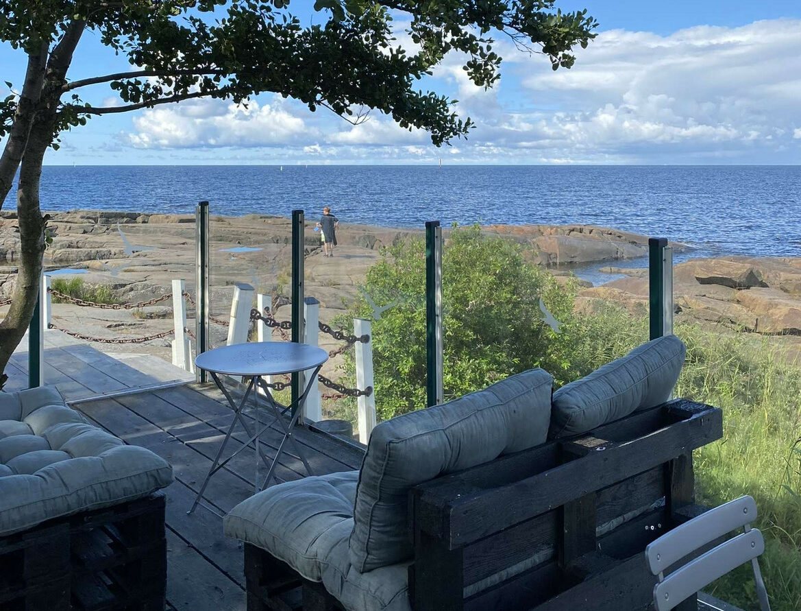 Café Kallon terassi: puiset sohvat, valkoiset tolpat ja köysikaide, taustalla avomeri